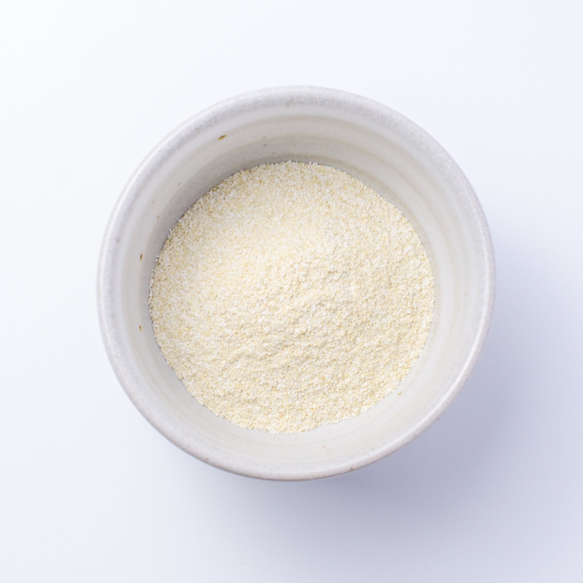 A top-down view of a small off-white ceramic bowl filled with Verdant Wild Brighten Facial Grains (chamomile & turmeric): a fine, pale yellow powder, set against a clean white background.