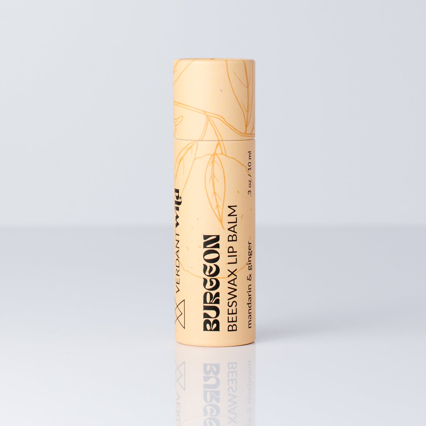 A single tube of Burgeon Beeswax Lip Balm in a pale peach-beige color with gold leaf illustrations and black text, standing upright on a reflective light gray surface.