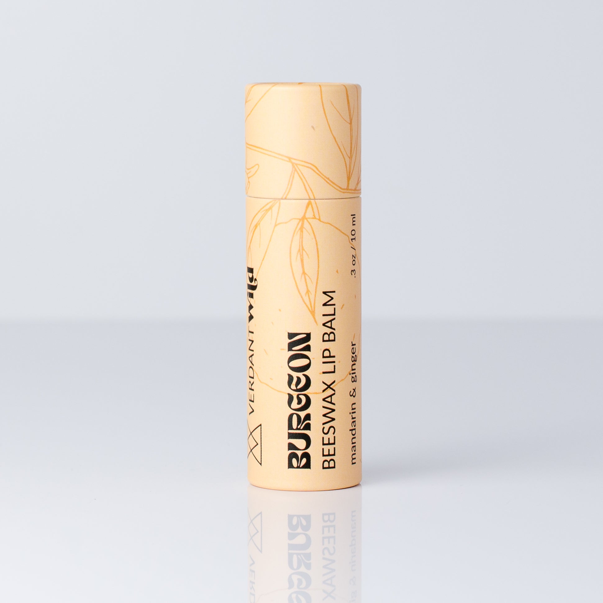 A single tube of Burgeon Beeswax Lip Balm in a pale peach-beige color with gold leaf illustrations and black text, standing upright on a reflective light gray surface.