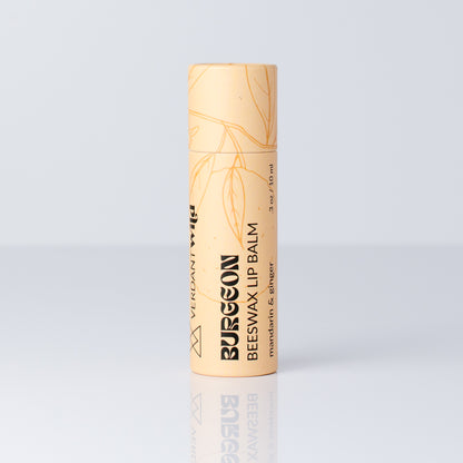 A single tube of Burgeon Beeswax Lip Balm in a pale peach-beige color with gold leaf illustrations and black text, standing upright on a reflective light gray surface.