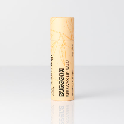 A single tube of Burgeon Beeswax Lip Balm with a pale peach-beige cap and body, featuring gold botanical line art and black text, set against a clean white background with a soft reflection below.