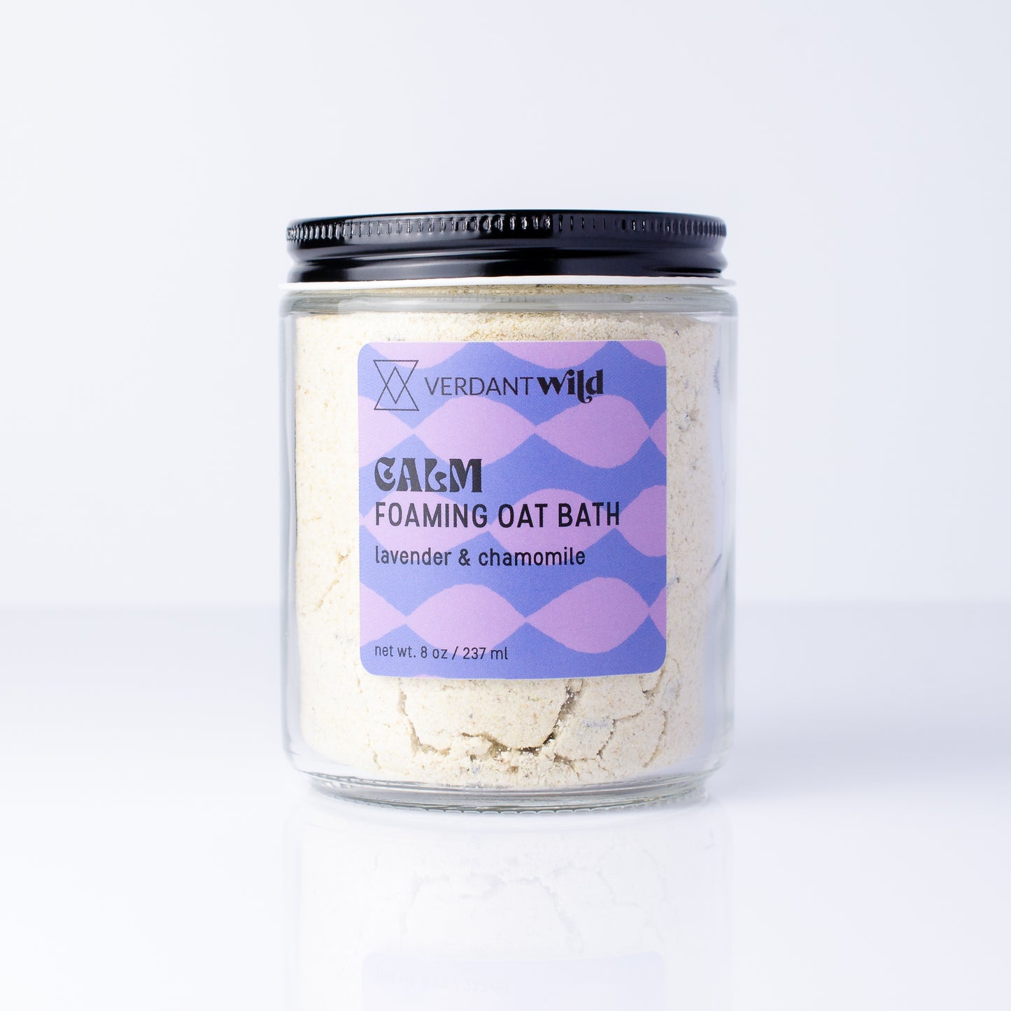 A clear glass jar of Verdant Wild Foaming Oat Bath in lavender and chamomile, with a black lid, set against a clean white background.