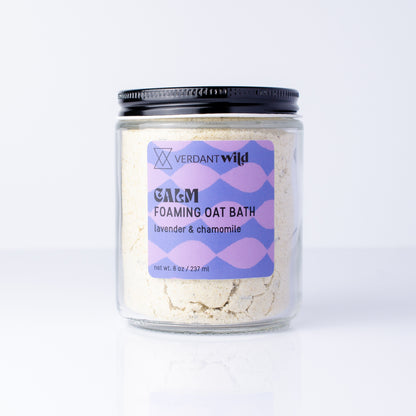 A clear glass jar of Verdant Wild Foaming Oat Bath in lavender and chamomile, with a black lid, set against a clean white background.