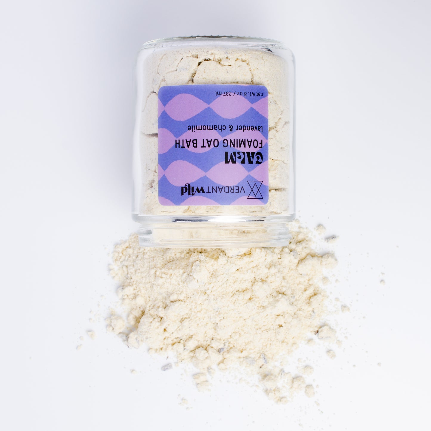 A clear glass jar filled with a powdered milk bath, featuring a purple and lavender patterned label. Some of the beige powder is spilled in a small pile in front of the jar on a clean white background.