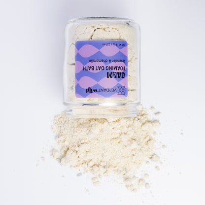 A clear glass jar filled with a powdered milk bath, featuring a purple and lavender patterned label. Some of the beige powder is spilled in a small pile in front of the jar on a clean white background.