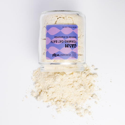A clear glass jar filled with a light beige powder, with some of the powder spilled in front on a white background. The jar has a purple and lavender label that reads 'VERDANT Wild' and 'FOAMING OAT BATH'.