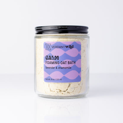 A clear glass jar of Verdant Wild Foaming Oat Bath in lavender & chamomile, filled with a light beige powder and topped with a black lid, set against a clean white background.