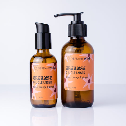 Two amber glass bottles of Verdant Wild Cleanse Oil Cleanser on a white reflective surface. One bottle is a smaller pump bottle and the other is a larger pump dispenser, both with black pumps and orange labels featuring pink floral accents.