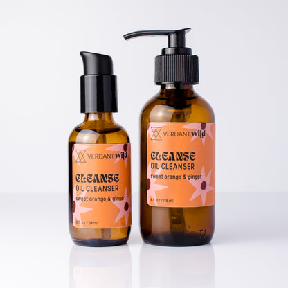 Two amber glass bottles of Verdant Wild Cleanse Oil Cleanser on a white reflective surface. One bottle is a smaller pump bottle and the other is a larger pump dispenser, both with black pumps and orange labels.
