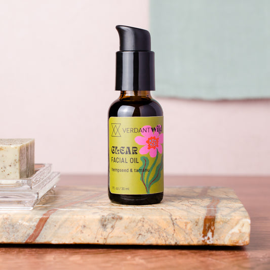 A bottle of Verdant Wild Clear Facial Oil with a black pump and a yellow-green label featuring a pink flower, displayed on a marble slab next to a bar of soap against a soft pink and mint background.