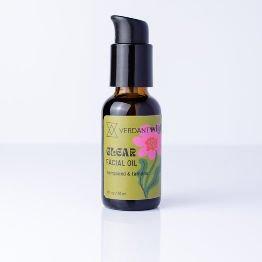 A 1 oz amber glass bottle of Verdant Wild Clear Facial Oil with a black pump dispenser and a yellow-green label featuring a pink flower, set against a clean white background.