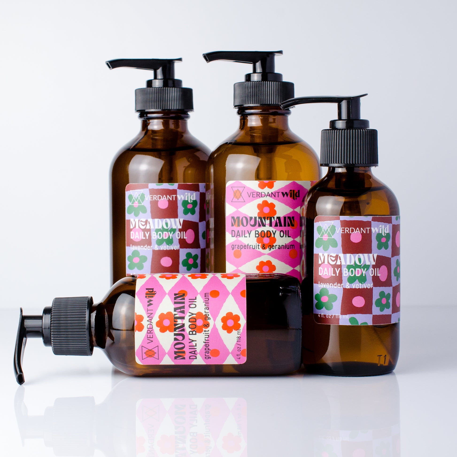Four amber glass pump bottles of Verdant Wild Daily Body Oil arranged on a white reflective surface, with one bottle lying down in front. The bottles have black pumps and colorful labels with pink, green, orange, and brown geometric patterns.