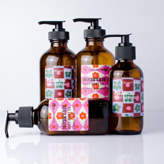 Four amber glass pump bottles of Verdant Wild Daily Body Oil arranged on a white reflective surface, with one bottle lying down in front. The bottles have black pumps and colorful labels with pink, green, orange, and brown geometric patterns.