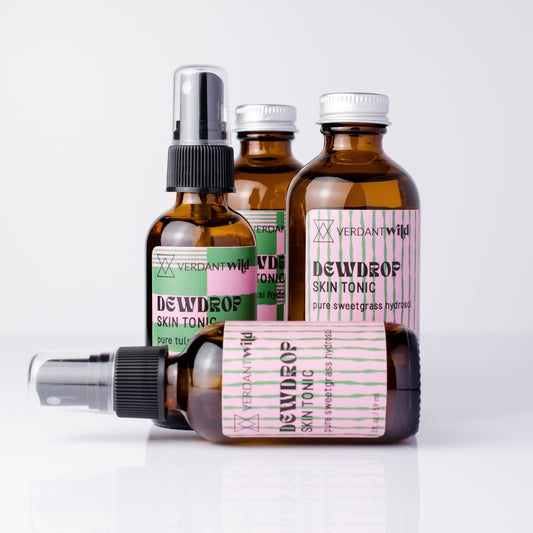 Four amber glass bottles of Verdant Wild Dewdrop Skin Tonic arranged on a white reflective surface against a light gray background.