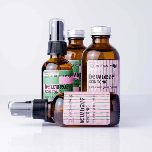 4 glass amber bottles of Verdant Wild Dewdrop Skin Tonic arranged on a white reflective surface against a light gray background, with one bottle lying down in front and 3 standing behind.