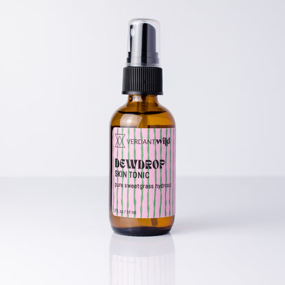 A 2 fl oz amber glass spray bottle of Verdant Dewdrop Skin Tonic with a black spray cap and a pink and white striped label, set against a clean white background with a soft reflection.