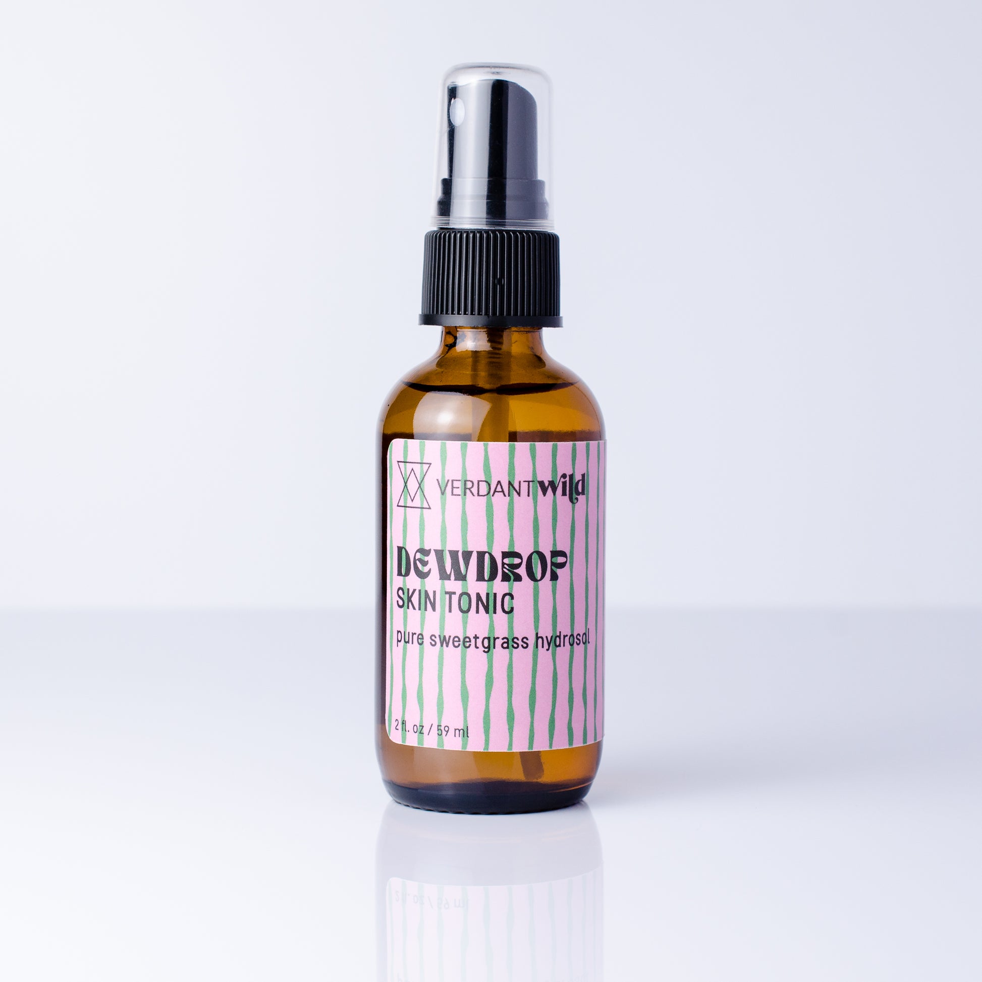 An amber glass spray bottle of Sweetgrass Skin Tonic with a black pump and a pink and white striped label, set against a clean white background with a soft reflection.