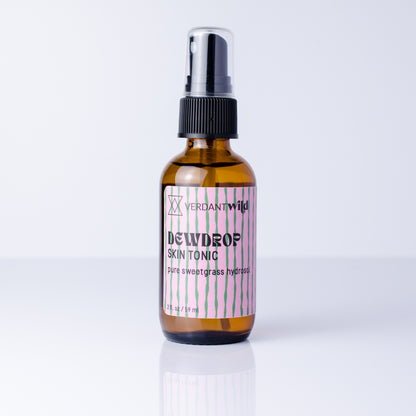 An amber glass spray bottle of Sweetgrass Skin Tonic with a black pump and a pink and white striped label, set against a clean white background with a soft reflection.