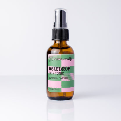 A 2 fl oz amber glass spray bottle of Verdant Wild Dewdrop Skin Tonic with a black spray cap and a colorful pink, green, and white label, set against a clean white background with a soft reflection.
