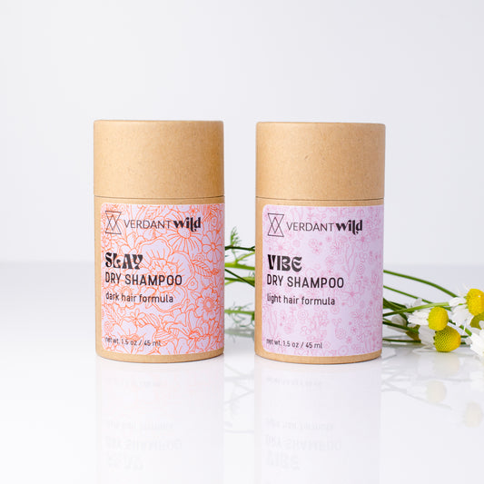 Two Verdant Wild dry shampoo tubes standing upright on a white reflective surface. The left tube is labeled Slay Dry Shampoo for dark hair, and the right tube is labeled Vibe Dry Shampoo for light hair.