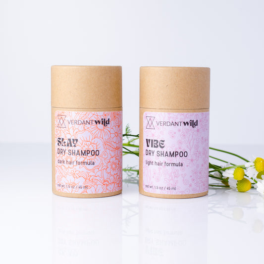 Two Verdant Wild dry shampoo tubes standing upright on a white reflective surface, with chamomile flowers placed to the right. The left tube is labeled 'Slay' with a red pattern and the right tube is labeled 'Vibe' with a pink pattern.