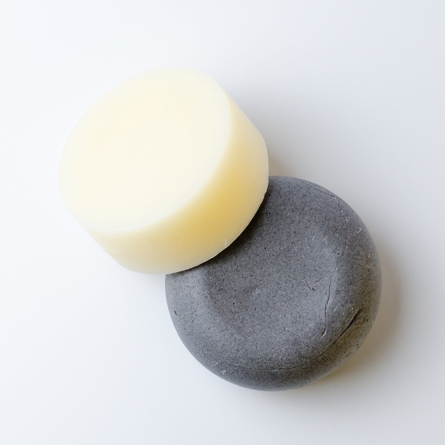 A shampoo and conditioner bar, one pale cream and one dark charcoal gray, arranged overlapping on a clean white background.