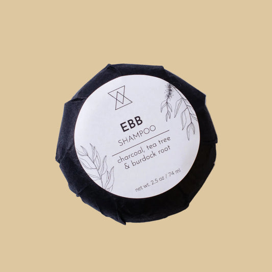 A round, dark charcoal-colored shampoo bar wrapped in black paper, featuring a white circular label with the text 'EBB SHAMPOO' and botanical illustrations, set against a plain background.