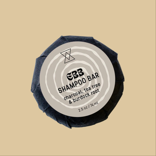A single wrapped shampoo bar on a clean background. The bar is wrapped in dark charcoal paper with a circular beige label reading 'charcoal, tea tree & burdock root'.
