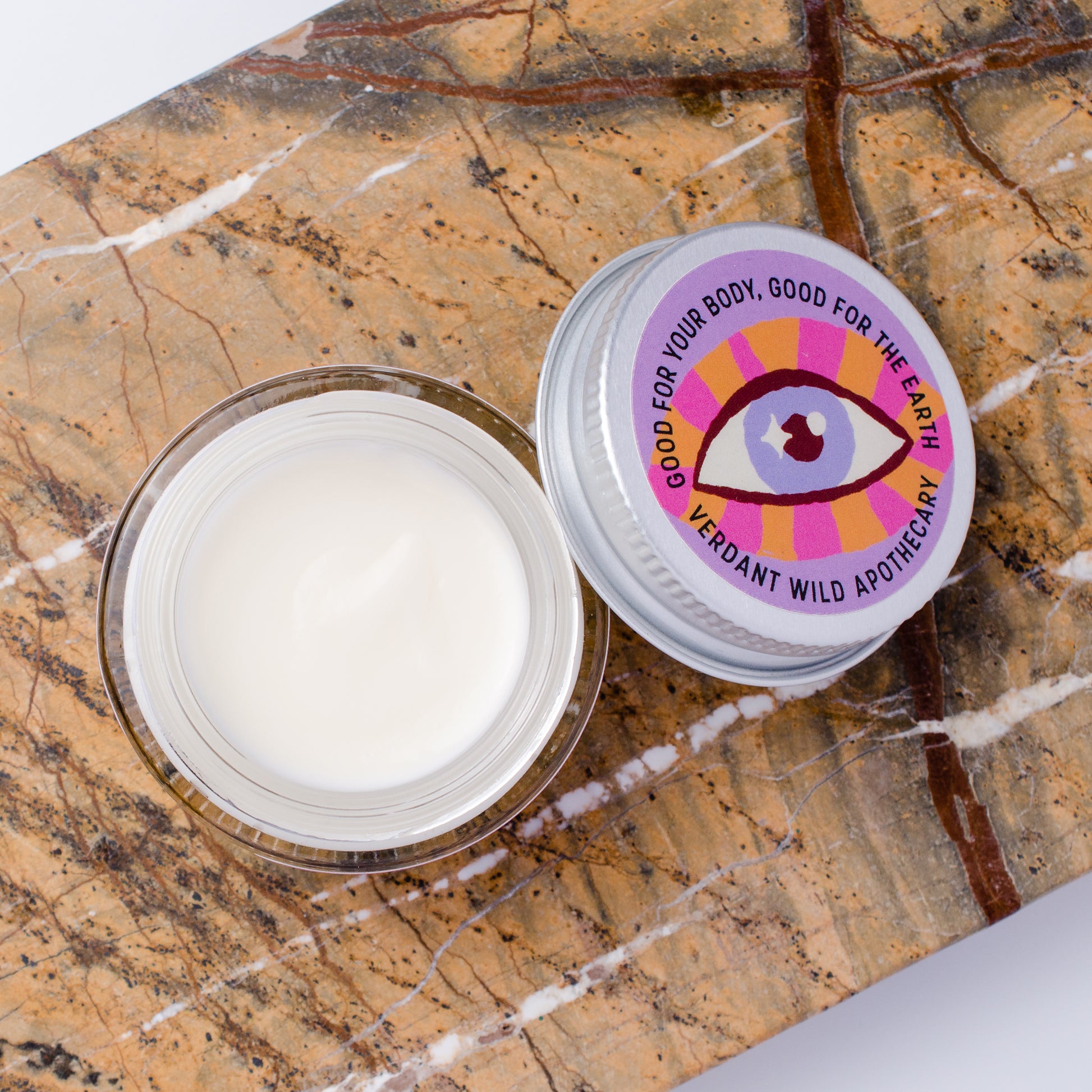 An open clear jar containing white cream sits beside a silver-lidded jar on a brown marble surface. The lid label shows a colorful eye graphic and the text 'GOOD FOR YOUR BODY, GOOD FOR THE EARTH' and 'VERDANT WILD APOTHECARY'.