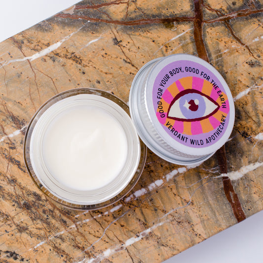An open clear jar containing white cream sits beside a silver-lidded jar on a brown marble surface. The lid label shows a colorful eye graphic and the text 'GOOD FOR YOUR BODY, GOOD FOR THE EARTH' and 'VERDANT WILD APOTHECARY'.