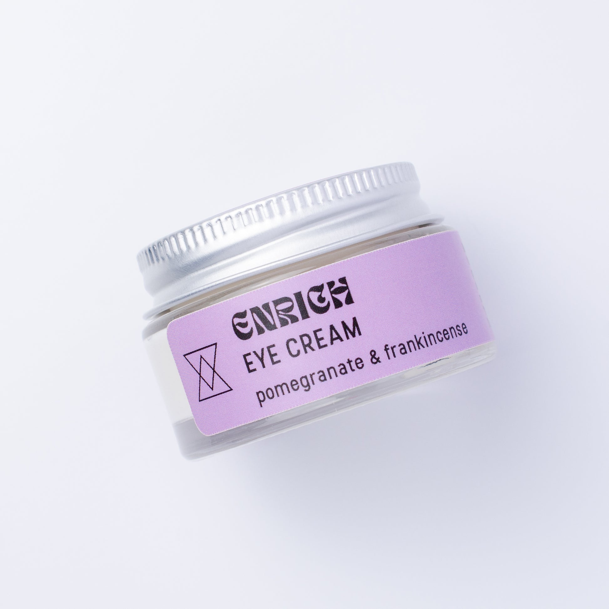 A small clear cosmetic jar of Enrich Eye Cream with a lavender label and silver lid, photographed on a clean white background.