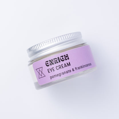 A small clear cosmetic jar of Enrich Eye Cream with a lavender label and silver lid, photographed on a clean white background.