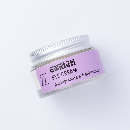 A small clear cosmetic jar of Enrich Eye Cream with a lavender label and silver lid, photographed on a clean white background.