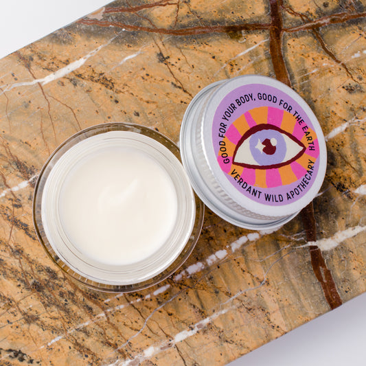 An open clear cosmetic jar filled with white cream sits beside its lid on a brown marble surface. The lid has a colorful circular label with an eye graphic and the text 'GOOD FOR YOUR BODY, GOOD FOR THE EARTH' and 'VERDANT WILD APOTHECARY'.