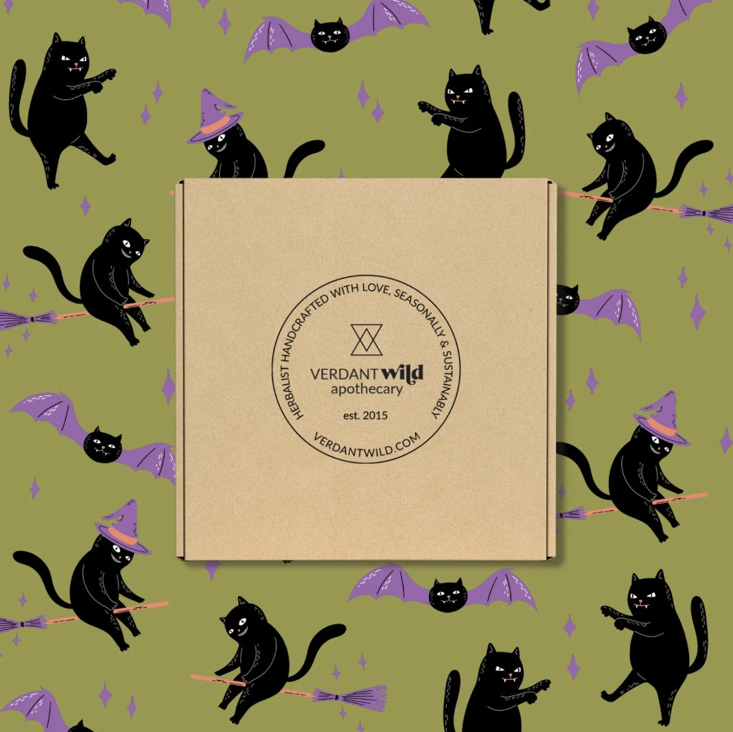 A square kraft paper box centered on a repeating Halloween-style pattern of black cats on broomsticks and purple bats. The box has a circular logo reading Verdant Wild Apothecary.
