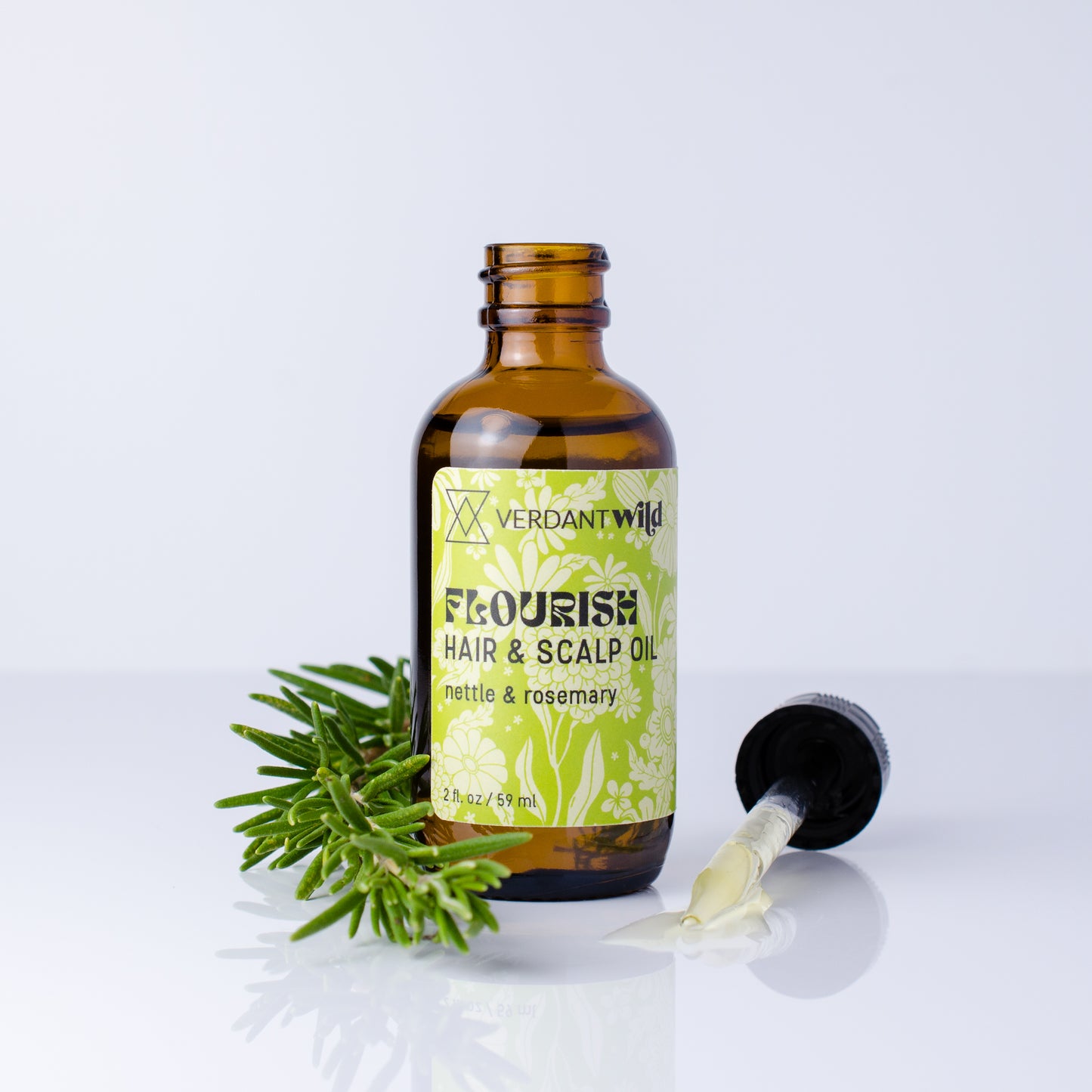 An amber glass bottle of Verdant Wild Flourish Hair & Scalp Oil with a green and white label, shown on a white reflective surface with a green rosemary sprig and a black dropper lying beside it.