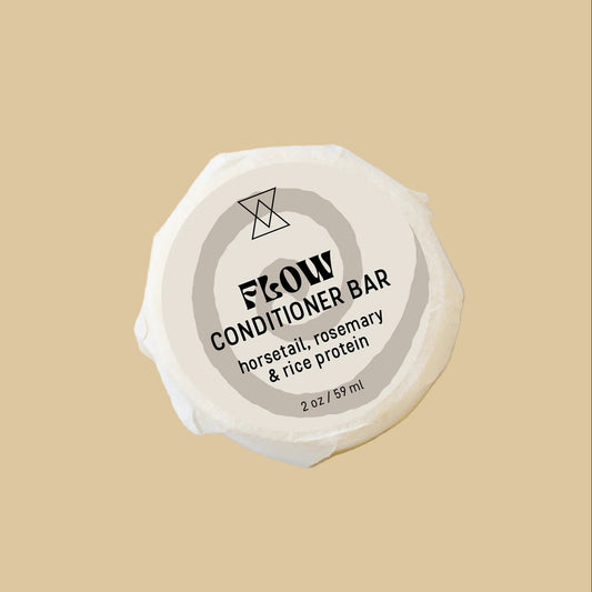 A round, unbleached soap bar wrapped in paper with a spiral label reading 'FLOW CONDITIONER' and 'horsetail, rosemary & rice protein' on a clean background.