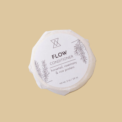 A round, unbleached soap bar wrapped in paper with a white label reading 'FLOW CONDITIONER' and 'horsetail, rosemary & rice protein' on a clean background.