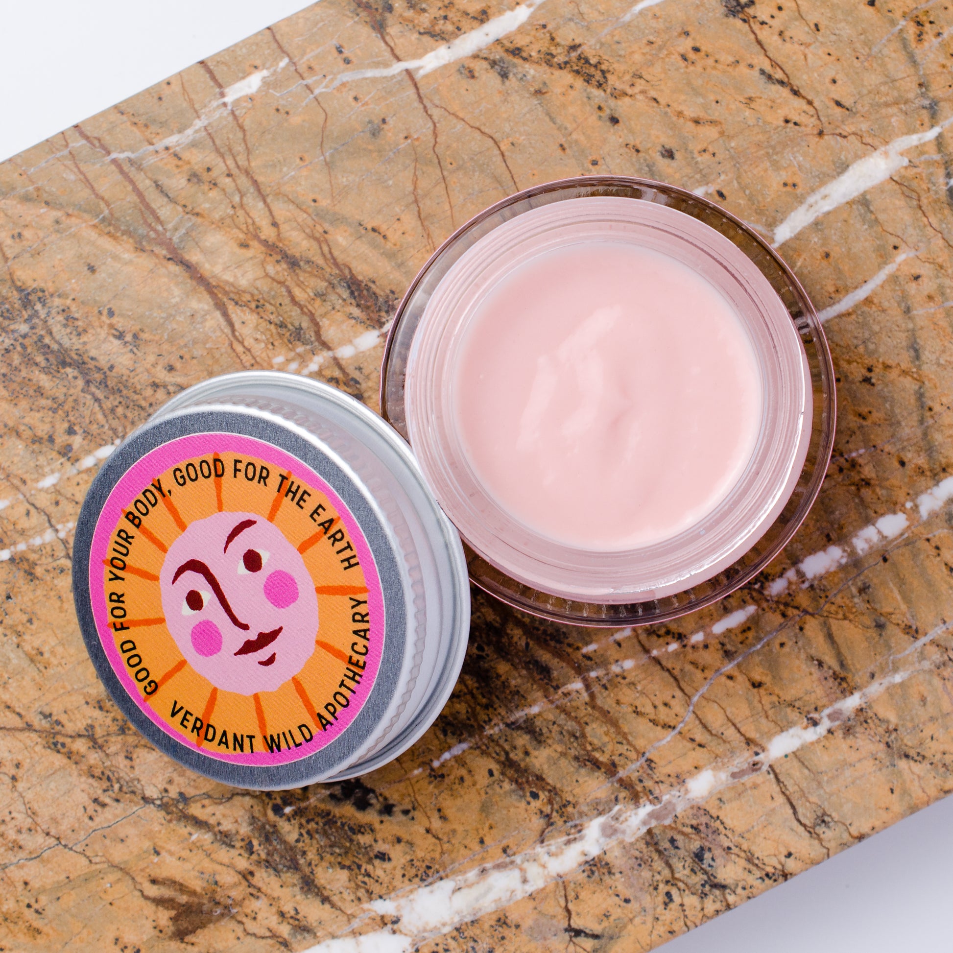 An open jar of pink cream next to its lid, which features a colorful circular label with a sun face and the text 'GOOD FOR YOUR BODY, GOOD FOR THE EARTH / VERDANT WILD APOTHECARY'. The products are styled on a brown and white veined marble surface.
