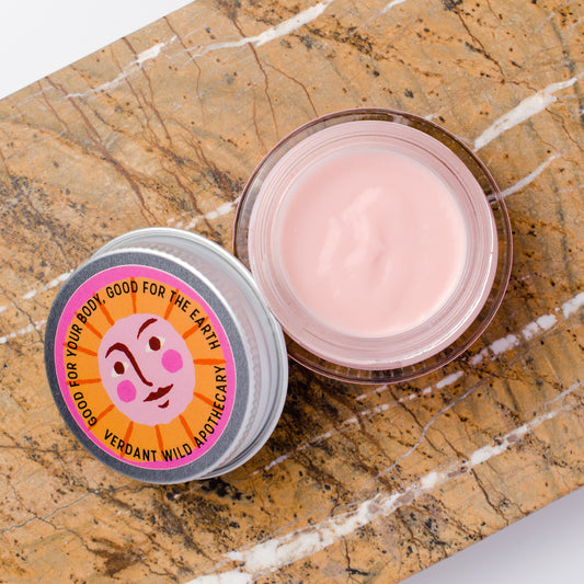 An open jar of pink face cream next to its lid, which features a colorful circular label with a cartoon face and the text 'GOOD FOR YOUR BODY, GOOD FOR THE EARTH / VERDANT WILD APOTHECARY'. The products are styled on a brown and white veined marble surface.