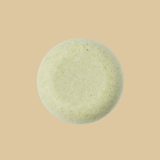 A single round, pale sage green shampoo bar with a textured, matte surface, photographed from directly above on a plain background.