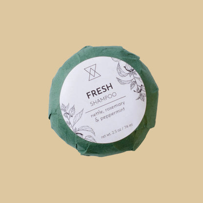 A round wrapped shampoo bar on a clean background. The product is wrapped in textured green paper with a white circular label reading 'FRESH SHAMPOO' and 'nettle, rosemary & peppermint'.
