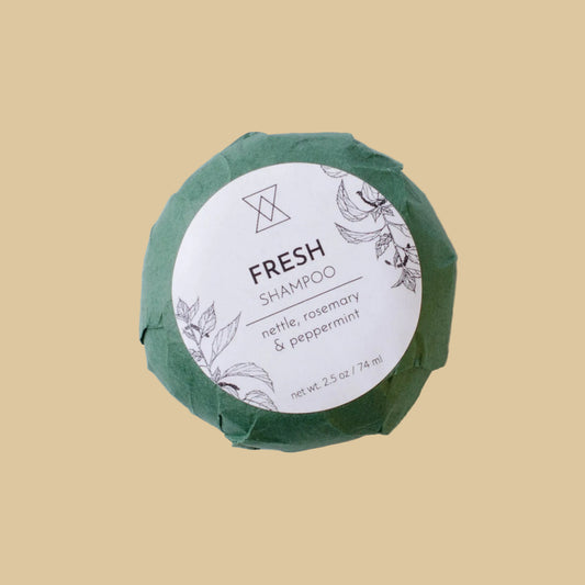 A round wrapped shampoo bar on a clean background. The product is wrapped in textured green paper with a white circular label reading 'FRESH SHAMPOO' and 'nettle, rosemary & peppermint'.