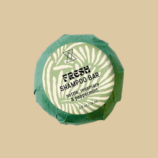A round wrapped shampoo bar on a clean background. The product is wrapped in textured green paper with a white circular label reading 'FRESH SHAMPOO' and 'nettle, rosemary & peppermint'.