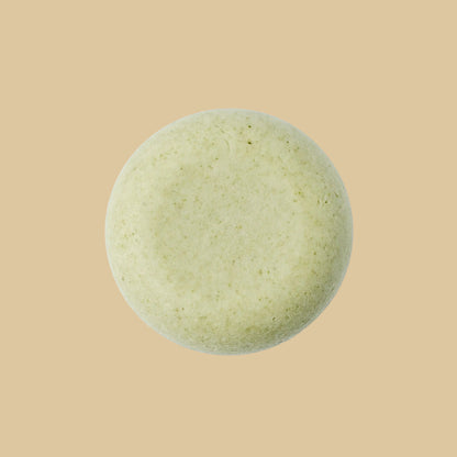 A single round, pale sage green shampoo bar with a textured, matte surface, photographed from directly above on a plain background.