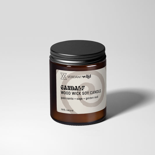 A glass jar candle with amber-brown wax and a black metal lid, featuring a white label with black text that reads 'GANDALF WOOD WICK SOY CANDLE'.