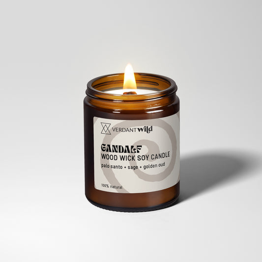 A lit amber glass jar candle with a grey label reading Verdant Wild GANDALF Wood Wick Soy Candle, photographed on a clean light gray studio background.