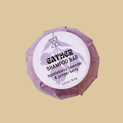 A single wrapped bar of shampoo on a clean background. The product is wrapped in a textured, faceted dusty purple paper wrapper with a white circular label reading 'GATHER SHAMPOO' and 'huckleberry, lavender & juniper berry'.