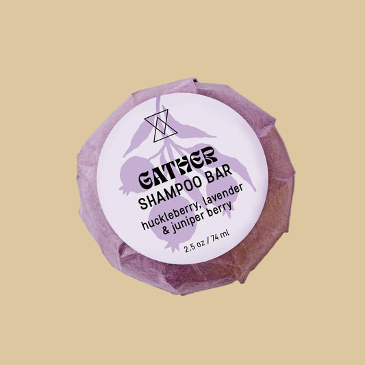 A single wrapped bar of shampoo on a clean background. The product is wrapped in a textured, faceted dusty purple paper wrapper with a white circular label reading 'GATHER SHAMPOO' and 'huckleberry, lavender & juniper berry'.