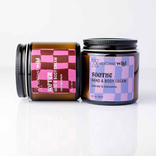 Two amber glass jars of hand and body cream on a white reflective surface. One jar is labeled 'SOFTEN' in pink and brown checkered packaging, and the other is labeled 'SOOthe' in purple and pink checkered packaging.